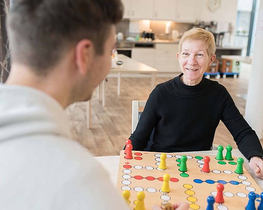Ältere Frau lächelt an einem Tisch beim Brettspiel; gegenüber sitzt ein jüngerer Mitspieler unscharf im Vordergrund, Spielbrett mit bunten Spielfiguren.