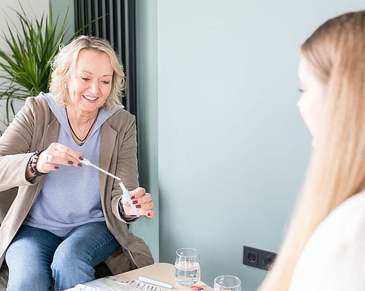 Frau demonstriert einer Patientin die Anwendung eines ISK-Katheters für Frauen; Gegenüber im Vordergrund unscharf