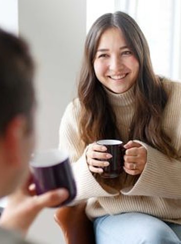 Zwei Personen unterhalten sich bei Kaffee; im Vordergrund unscharfe Person, im Fokus eine lächelnde Frau mit Tasse in heller Umgebung.
