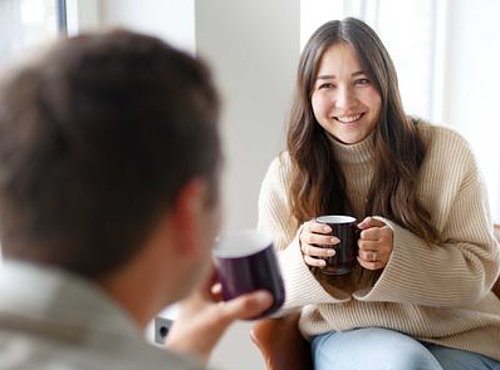 Zwei Personen unterhalten sich bei Kaffee; im Vordergrund unscharfe Person, im Fokus eine lächelnde Frau mit Tasse in heller Umgebung.