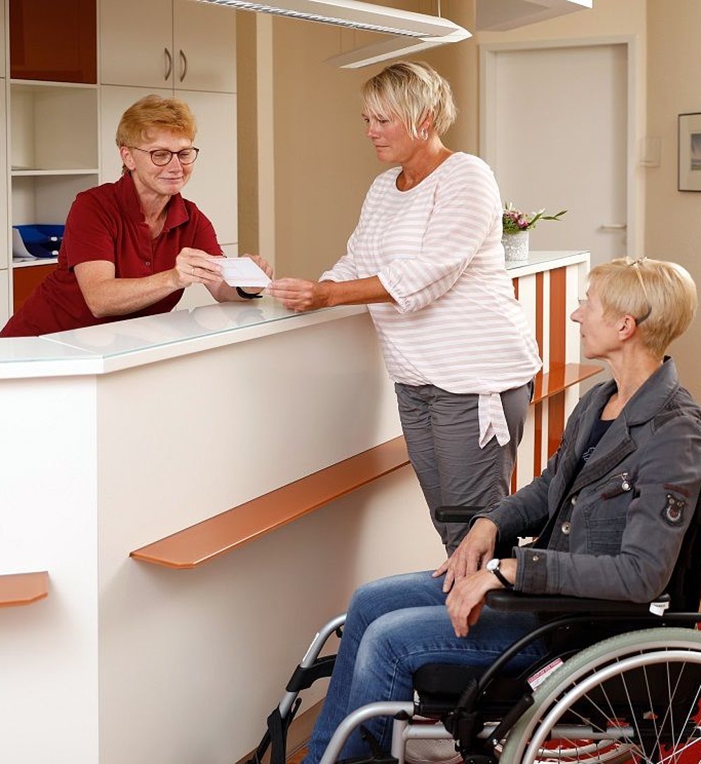 Eine Mitarbeiterin an der Rezeption begrüßt eine Besucherin, während eine weitere Frau im Rollstuhl daneben wartet. Die Szene wirkt freundlich und professionell und zeigt eine barrierefreie Empfangssituation im medizinischen oder sozialen Umfeld.