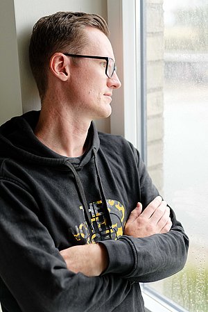 Ein Mann steht am Fenster und blickt nach draußen. Mit verschränkten Armen und nachdenklicher Haltung wirkt die Szene ruhig und introspektiv.