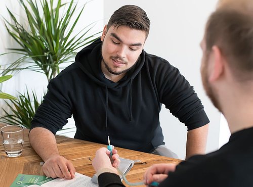 Zwei Männer im Beratungsgespräch am Tisch; einer zeigt mit einem Stift auf Unterlagen, der andere hört aufmerksam zu. Ein Katheter wird erklärt