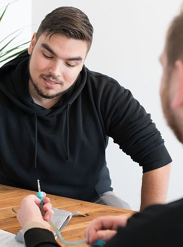 Zwei Männer im Beratungsgespräch am Tisch; einer zeigt mit einem Stift auf Unterlagen, der andere hört aufmerksam zu. Ein Katheter wird erklärt