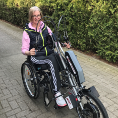 „Person im Rollstuhl mit Handbike-Vorsatz fährt auf einem gepflasterten Weg entlang einer Hecke; sportliche Kleidung, Brille, lächelt in die Kamera. Am Handbike ist ein Akkugehäuse sichtbar