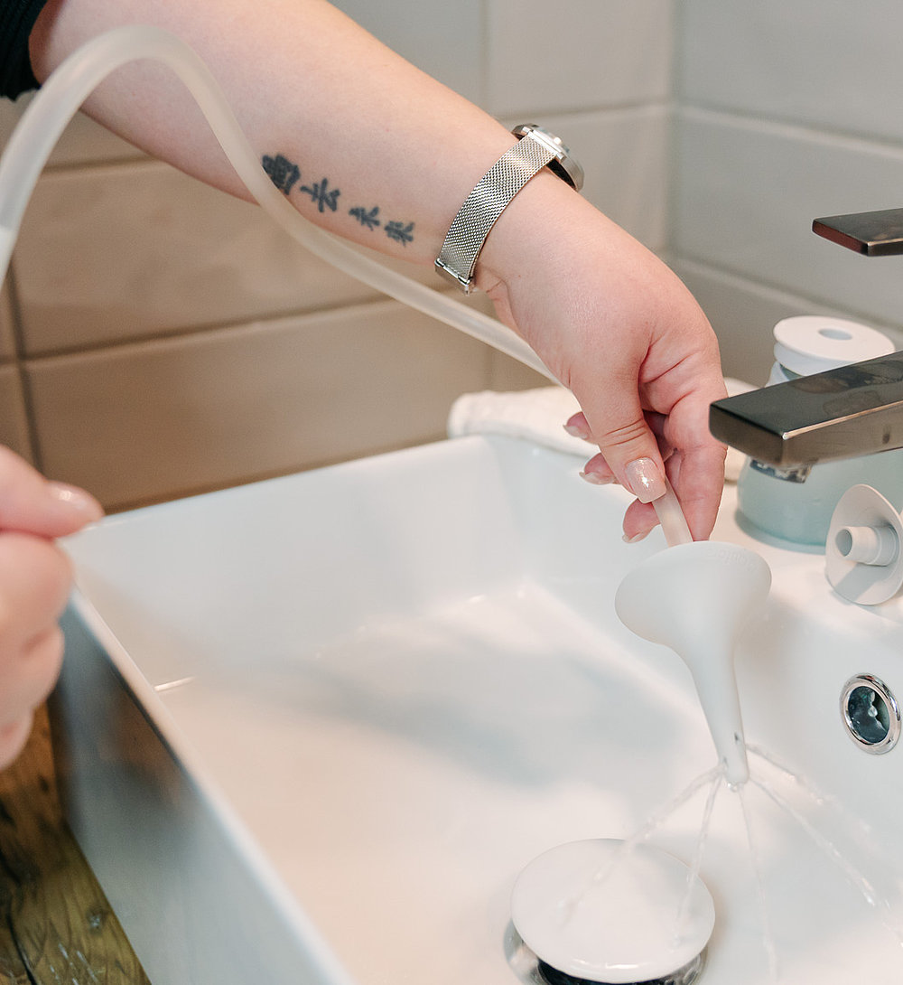 Nahaufnahme einer Person, die an einem Waschbecken den Schlauch eines Darm-Irrigationssystems mit Wasser füllt; neben dem Wasserhahn steht weiteres Zubehör bereit.