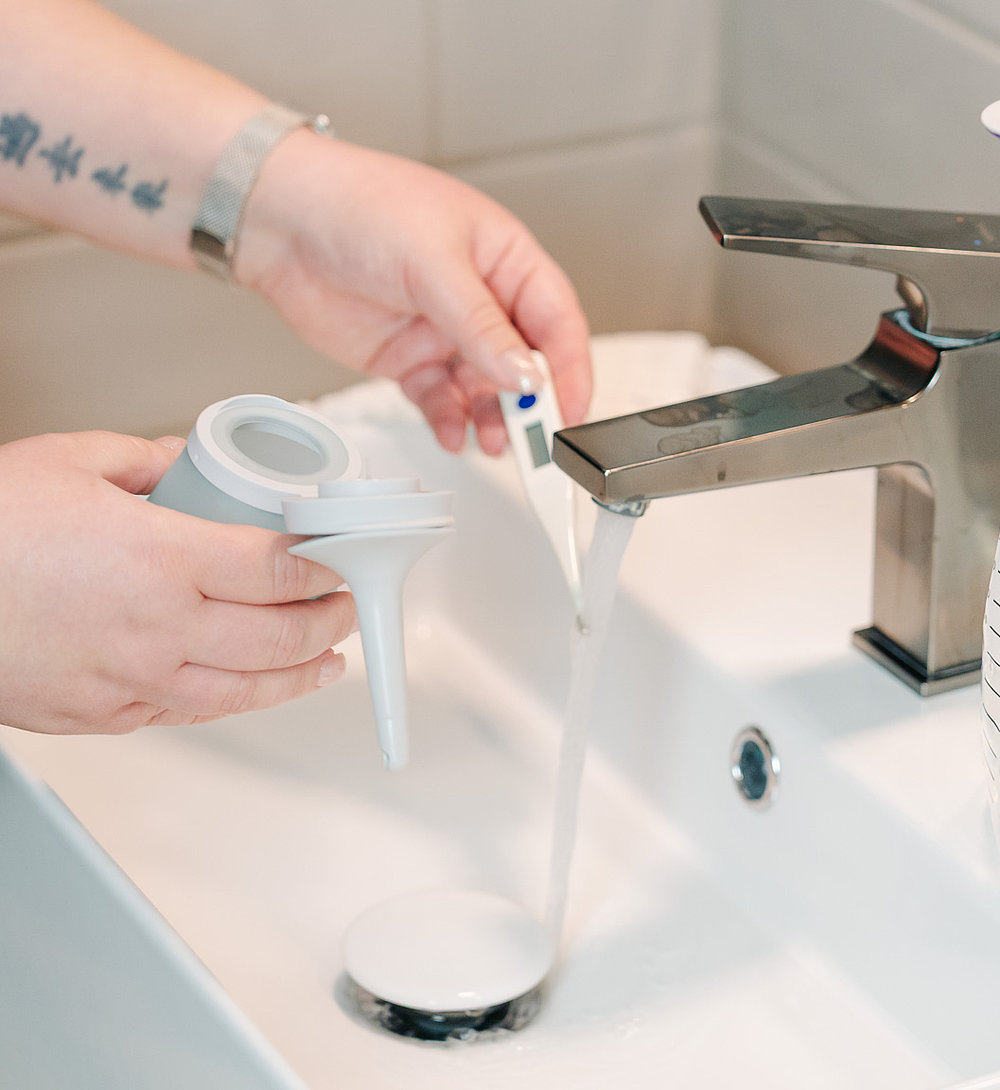Nahaufnahme einer Person, die am Waschbecken ein Trichterteil eines Darm-Irrigationssystems unter fließendem Wasser reinigt; daneben hängt ein mit Skala versehenes Irrigationsbeutel-System am Wasserhahn.