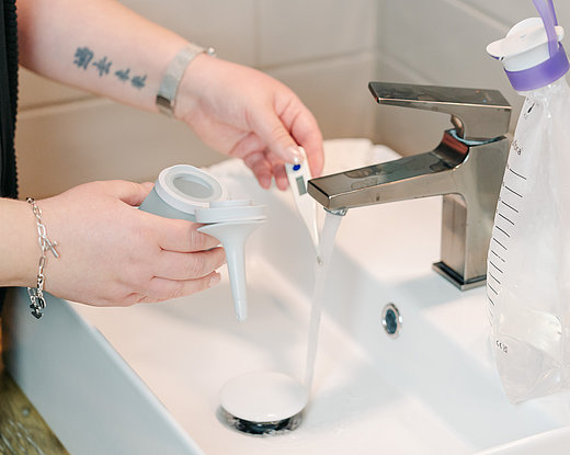 Nahaufnahme einer Person, die am Waschbecken den Trichter eines Darm-Irrigationssystems unter fließendem Wasser hält; daneben hängt ein transparenter Irrigationsbeutel mit Skala am Wasserhahn.