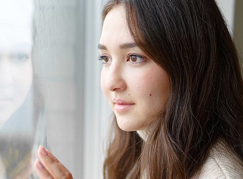 Frau im cremefarbenen Strickpullover blickt nachdenklich aus einem hellen Fenster und berührt die Scheibe.