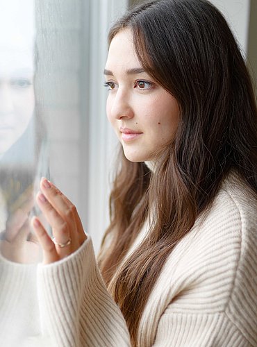 Frau im cremefarbenen Strickpullover blickt nachdenklich aus einem hellen Fenster und berührt die Scheibe.