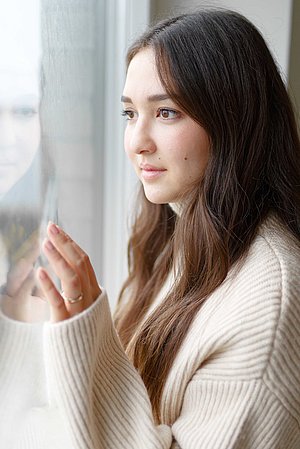 Eine junge Frau steht am Fenster und blickt nachdenklich nach draußen. Ihre Hand berührt leicht das Glas, während sie ruhig und in Gedanken versunken wirkt.