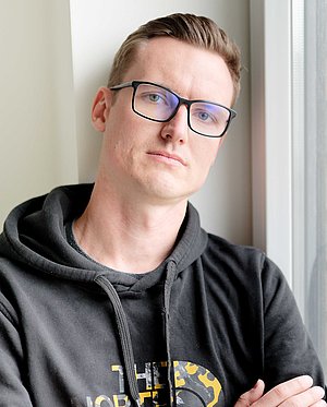 Ein Mann steht mit verschränkten Armen am Fenster und blickt ernst oder nachdenklich in die Kamera. Die Szene wirkt ruhig und reflektiert.