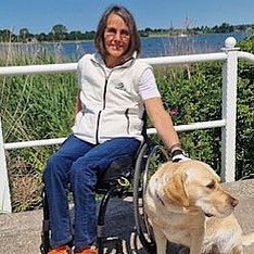 Person im Rollstuhl sitzt an einem Geländer am See; neben ihr sitzt ein heller Hund. Sonniger Tag, Schilf und Wasser im Hintergrund.