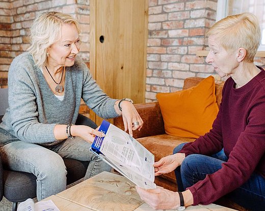 Zwei Frauen sitzen einander zugewandt auf einem Sofa und besprechen Broschüren; gemütliche Wohnumgebung mit Backsteinwand und Kissen.