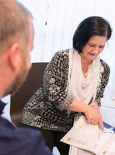 Frau führt mit einem Mann an einem Schreibtisch ein Gespräch und erklärt ihm ein Dokument.