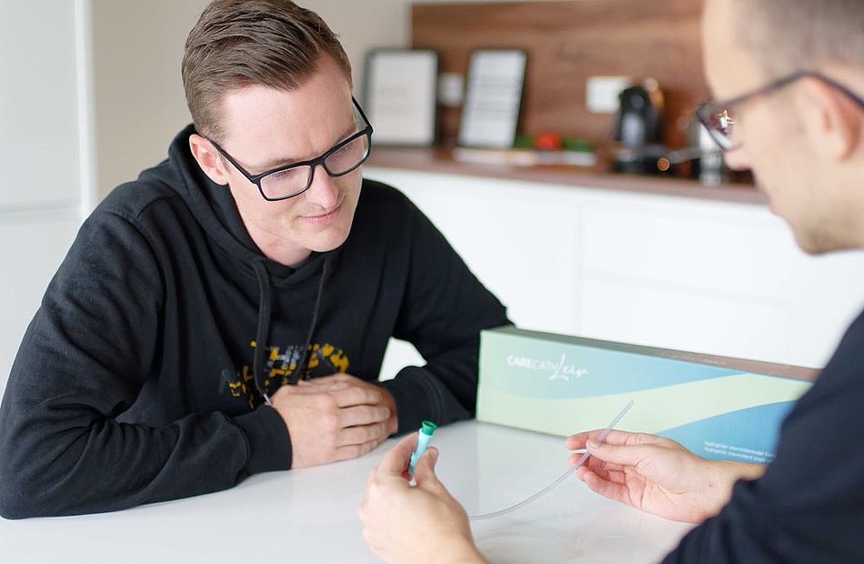 Zwei Männer am Tisch; ein Berater zeigt einen ISK-Einmalkatheter mit grünem Anschluss, der andere hört aufmerksam zu.