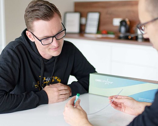 Zwei Männer am Tisch; ein Berater zeigt einen ISK-Einmalkatheter mit grünem Anschluss, der andere hört aufmerksam zu.