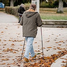Von hinten gesehene Person auf einem herbstlichen Gehweg; sie nutzt zwei Gehstützen und trägt Jacke, Jeans und Stiefel. Trockene Blätter liegen auf dem Boden.